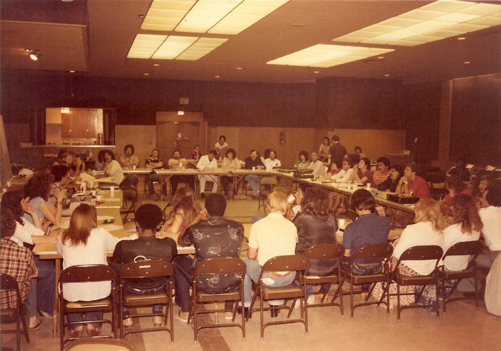 Group of people sitting around tables that are in the shape of a square. They're in what looks like an old community room.